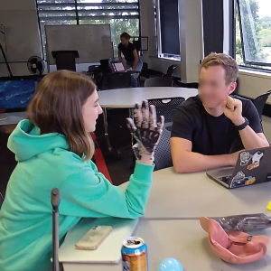 A team member wearing vinyl gloves with drawn-on joints demonstrates the hand gestures enabled by the prototype to the user, who observes attentively. The setting is a classroom workspace with laptops, props, and other prototype components on the table.